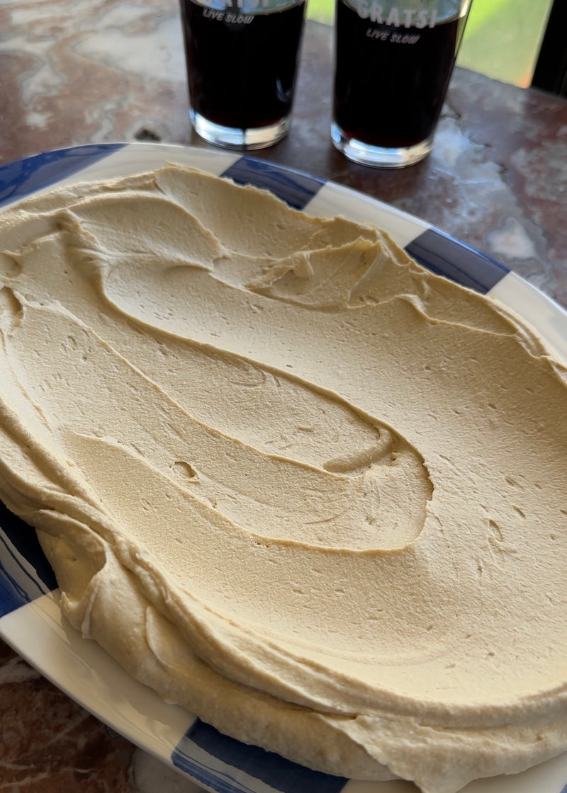 A blue and white plate holds a smooth, thick layer of light brown whipped dip or spread. In the background, two dark drinks in glasses sit on a marble surface near a window.