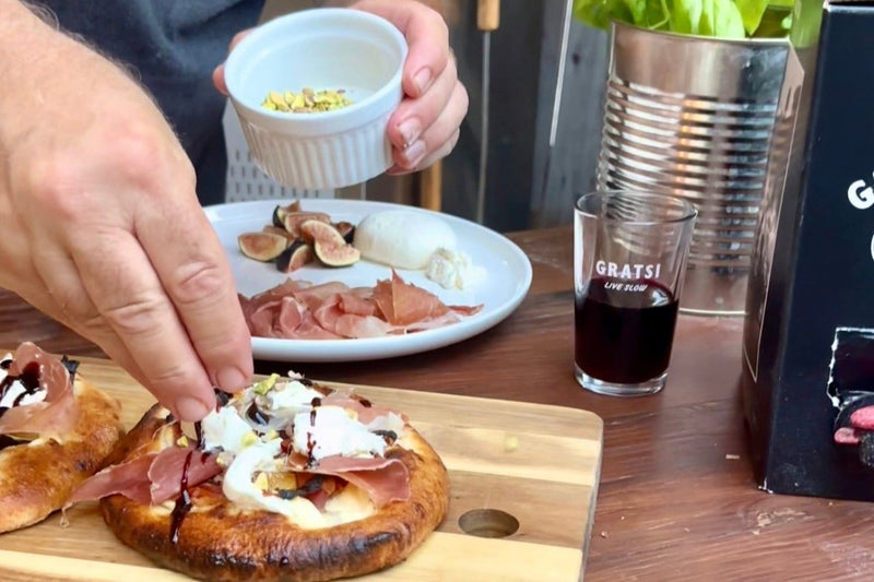 A person sprinkles chopped nuts onto a small pizza topped with prosciutto, cheese, and balsamic glaze. Nearby are a plate with figs, mozzarella, and prosciutto, a glass of red wine, and a basil plant in a tin pot.