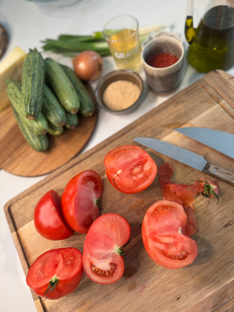Halved tomatoes on a wooden cutting board with a knife; in the background are whole cucumbers, an onion, a glass of liquid, spices, ketchup, olive oil, and cheese on a counter.