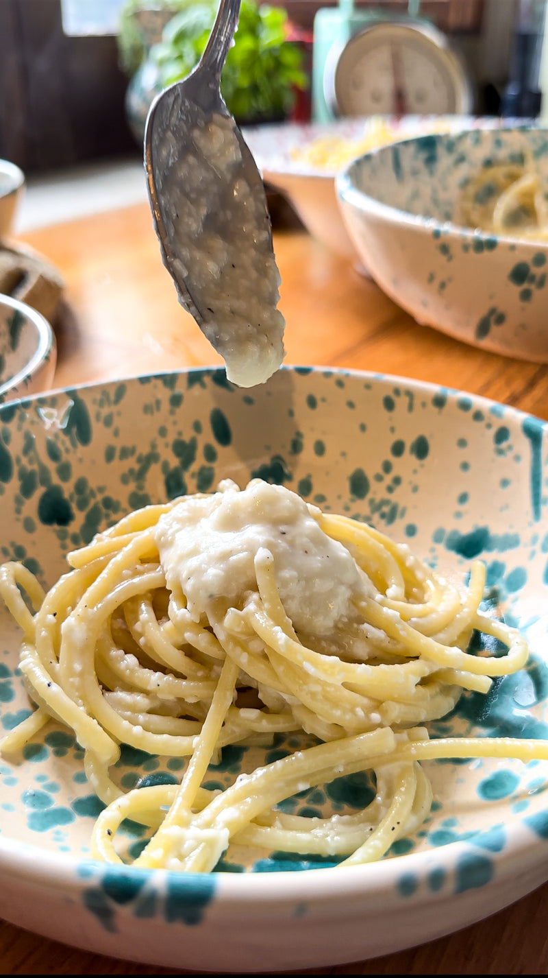 A close-up of spaghetti on a patterned ceramic plate as a spoon adds a creamy white sauce on top. The scene is set on a wooden table with blurred bowls and kitchen items in the background.