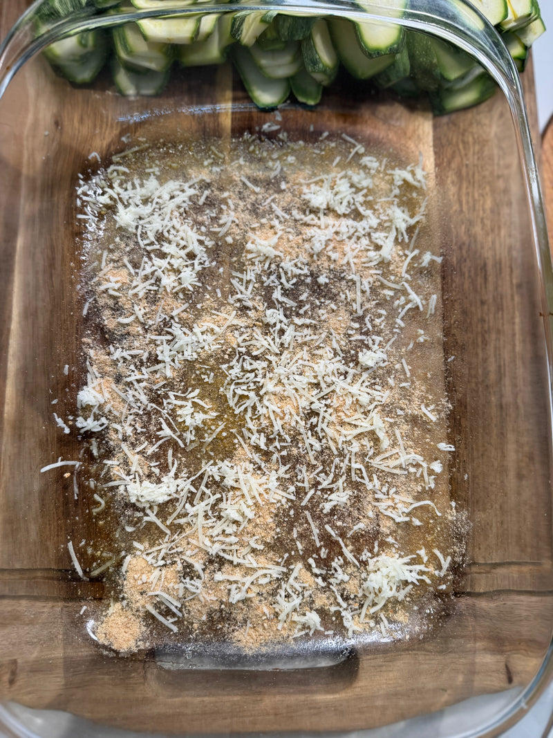 A glass baking dish contains a mixture of oil, spices, and shredded cheese, with sliced zucchini stacked in the background on a wooden surface.
