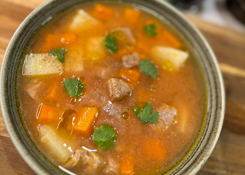 A bowl of hearty soup containing chunks of beef, carrots, potatoes, and garnished with fresh cilantro, served in a green-rimmed bowl on a wooden surface.