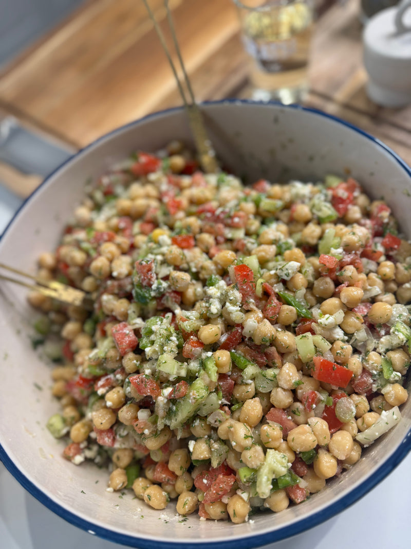 A close-up of a large bowl filled with chickpea salad mixed with chopped red bell peppers, cucumbers, herbs, and crumbled feta cheese, with a glass of water and wooden utensils in the background.