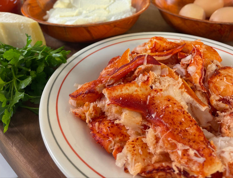 A plate of cooked lobster meat sits on a table next to fresh parsley, a wedge of cheese, eggs, and a bowl of ricotta cheese.