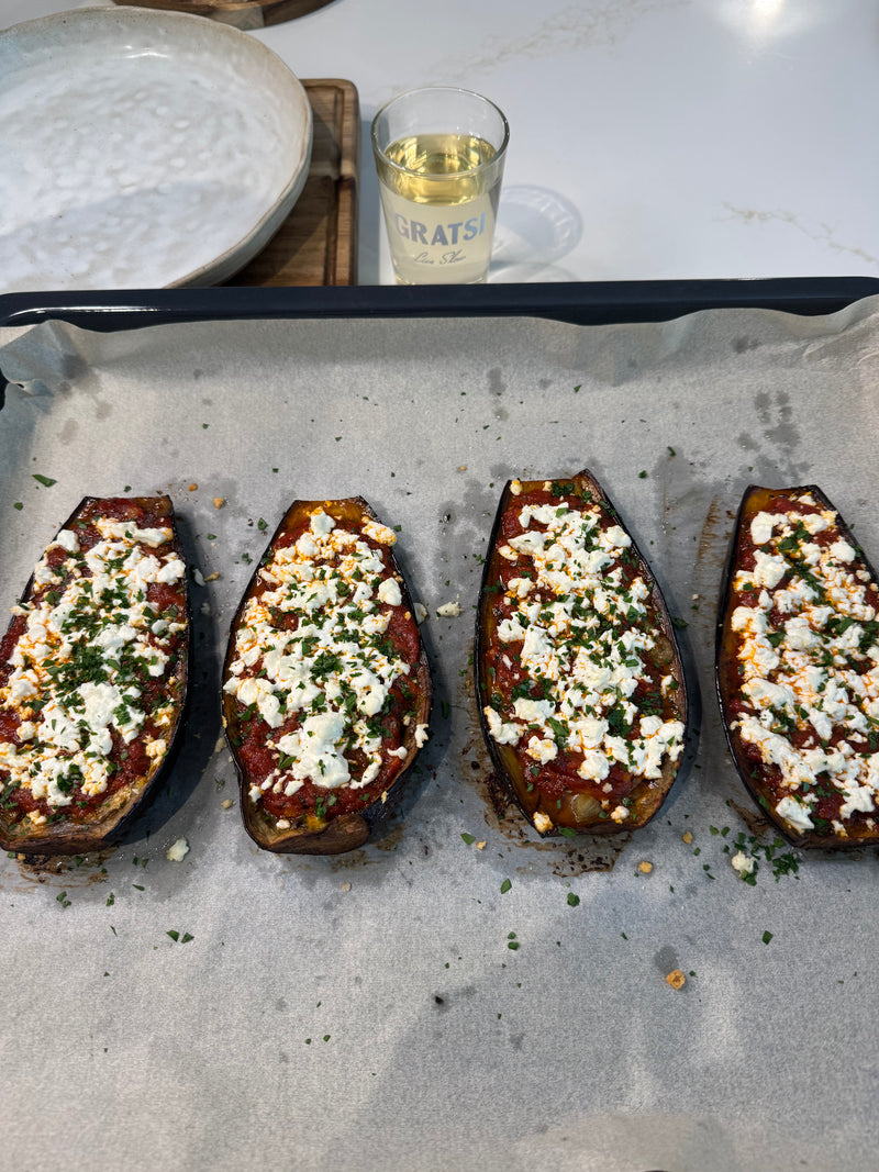 Four baked eggplant halves topped with tomato sauce, crumbled cheese, and chopped herbs on a parchment-lined baking tray. A plate and a glass of white wine are in the background.