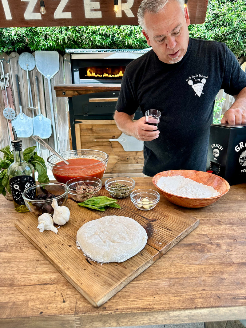 A man in a black T-shirt stands at an outdoor pizza station with pizza dough, flour, tomato sauce, fresh basil, olives, garlic, anchovies, and olive oil on a wooden table, with a wood-fired oven in the background.