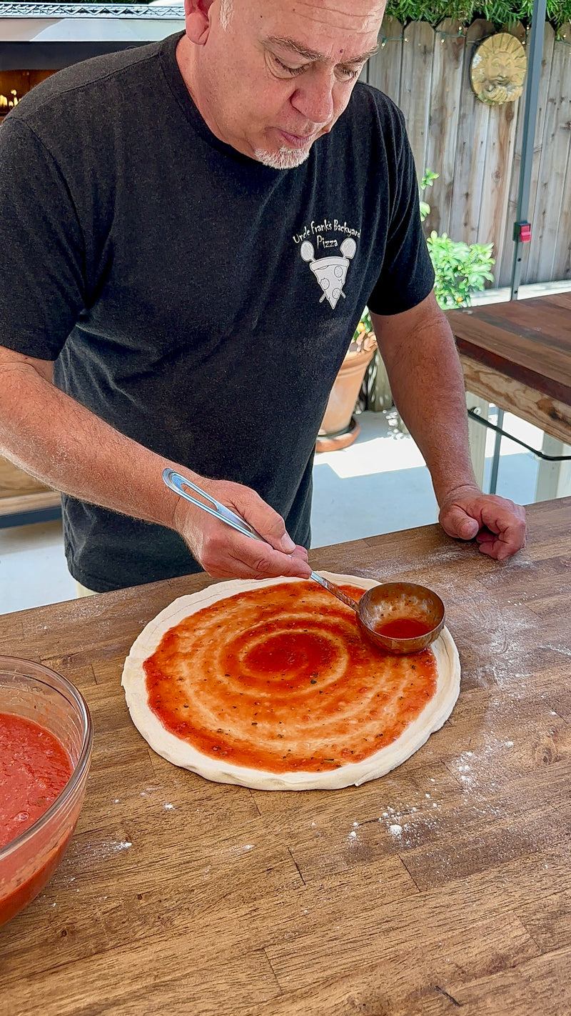 A man spreads tomato sauce on pizza dough with a ladle at a wooden table. A bowl of sauce sits nearby, and he wears a black t-shirt with a white pizza graphic. The setting appears to be outdoors.