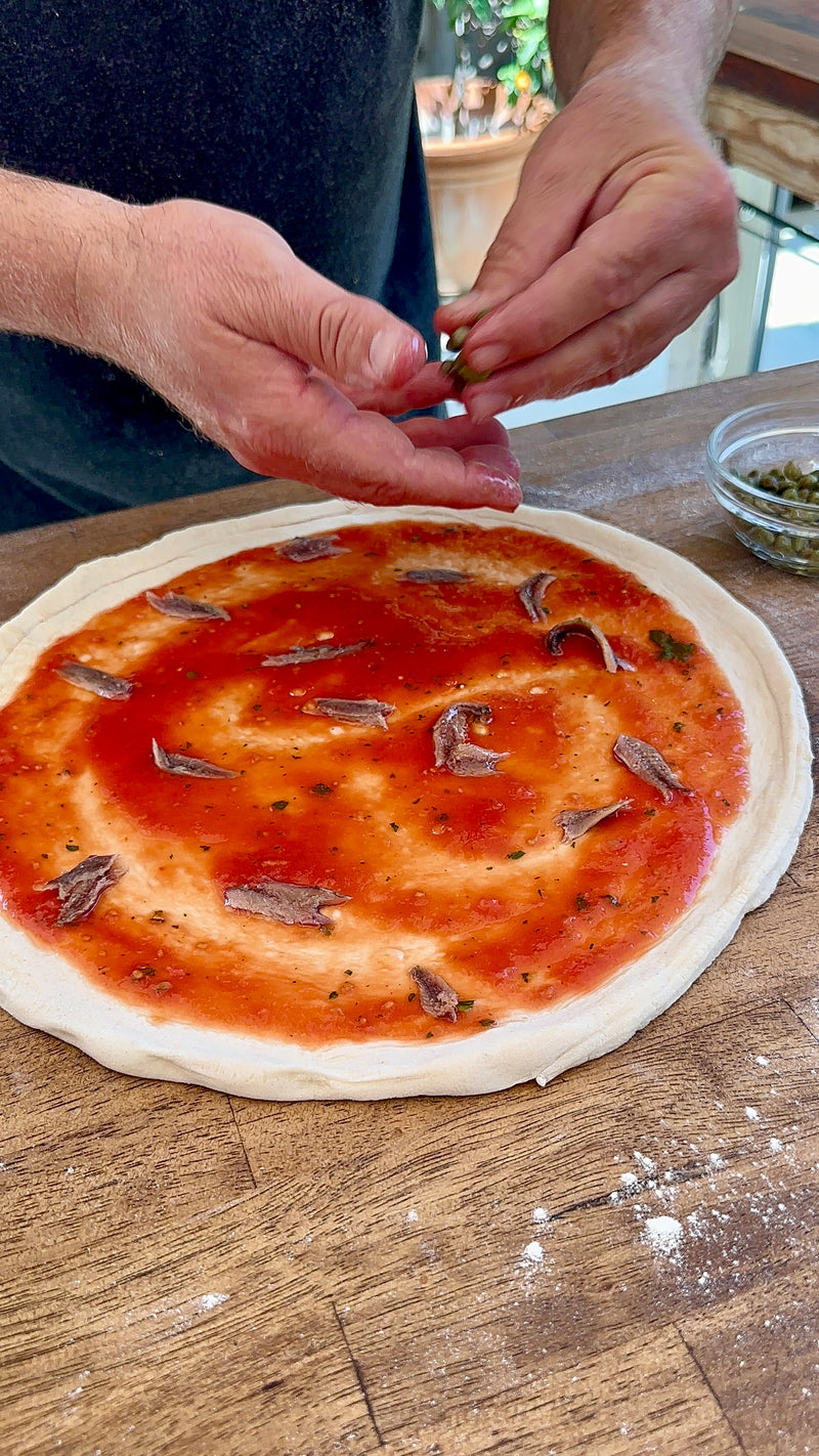 A person prepares a pizza by adding toppings to a pizza dough spread with tomato sauce and anchovy fillets on a wooden countertop.