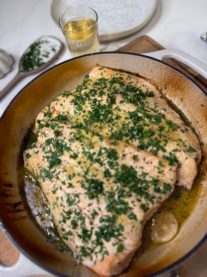 A large, cooked salmon fillet in a pan is topped with chopped herbs. A wooden spoon with more herbs, a glass of white wine, and a plate are visible in the background on a kitchen counter.