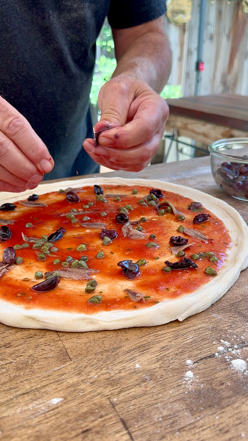 A person’s hands topping an uncooked pizza with black olives, capers, and anchovies on tomato sauce. The pizza dough is on a wooden surface, with a bowl of olives nearby. The background shows a bright, rustic kitchen.