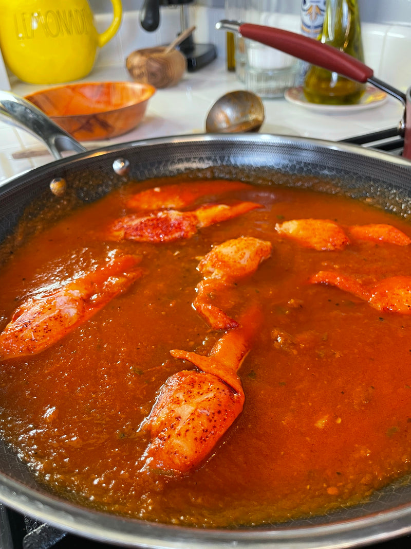A frying pan on a stove filled with crab claws simmering in a thick, red tomato-based sauce. Kitchen items like olive oil, a yellow container, and a ladle are visible in the background.