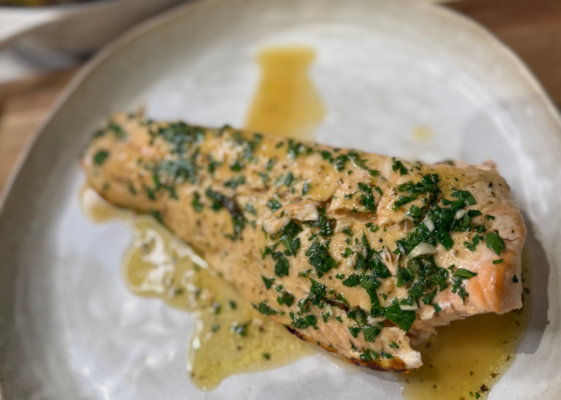 A cooked salmon fillet garnished with chopped herbs and drizzled with a light buttery sauce, served on an off-white plate.
