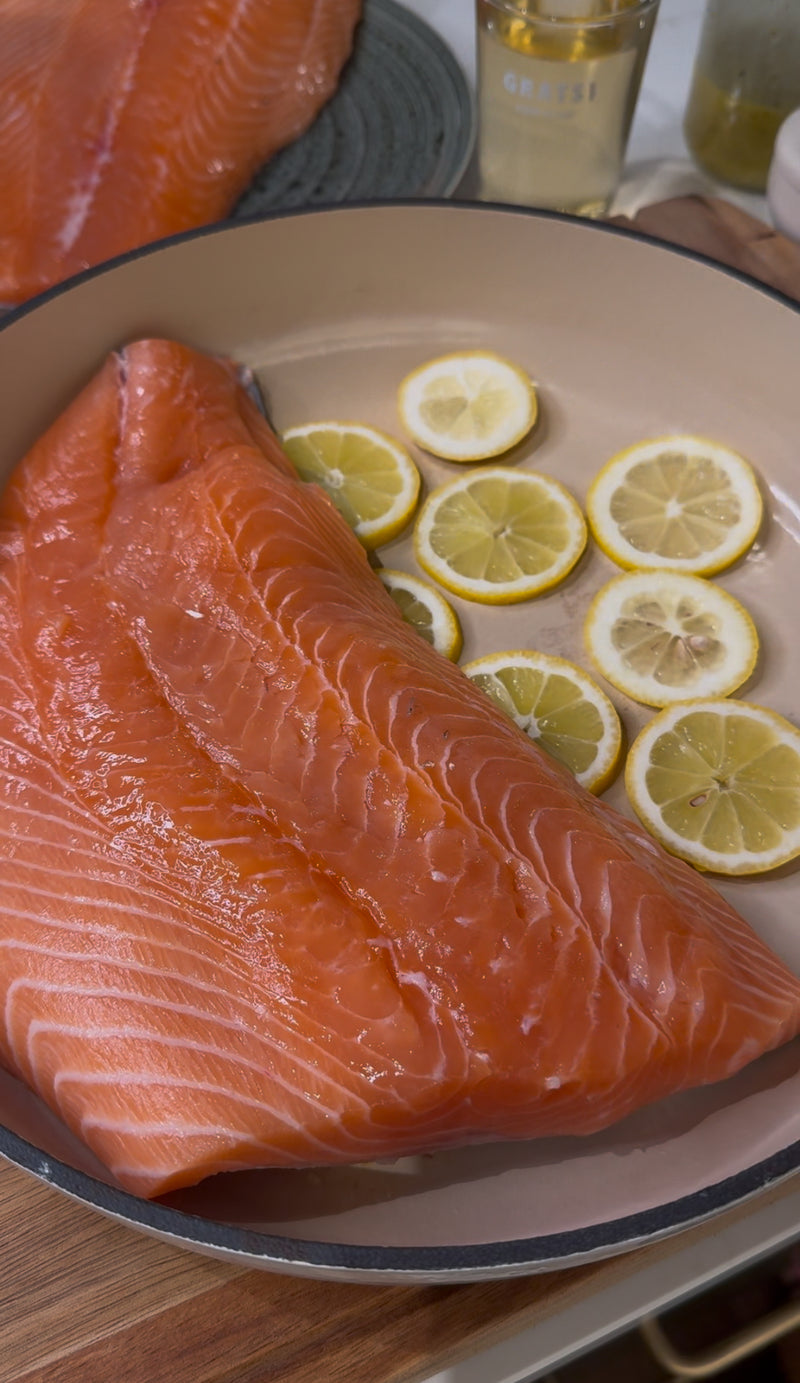 A large, raw salmon fillet rests on top of several thin lemon slices in a beige pan, ready to be cooked. A bottle of oil and a pepper grinder are partially visible in the background.