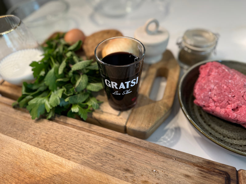 A glass with GRATSI written on it, filled with red wine, sits on a wooden board surrounded by parsley, an egg, a slice of bread, jars of spices, a bowl of ground beef, and a glass of milk.