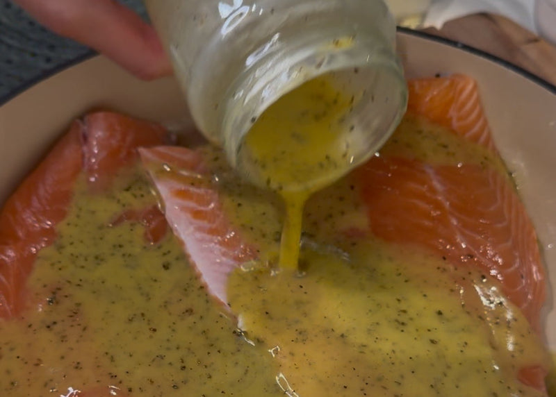 A hand pours a yellow, herb-speckled marinade from a jar onto raw salmon fillets in a dish, preparing them for cooking.