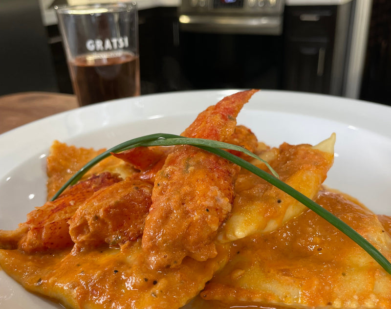 A close-up of ravioli topped with lobster meat and creamy tomato sauce, garnished with a green herb. A glass of rosé wine sits in the background on a kitchen counter.