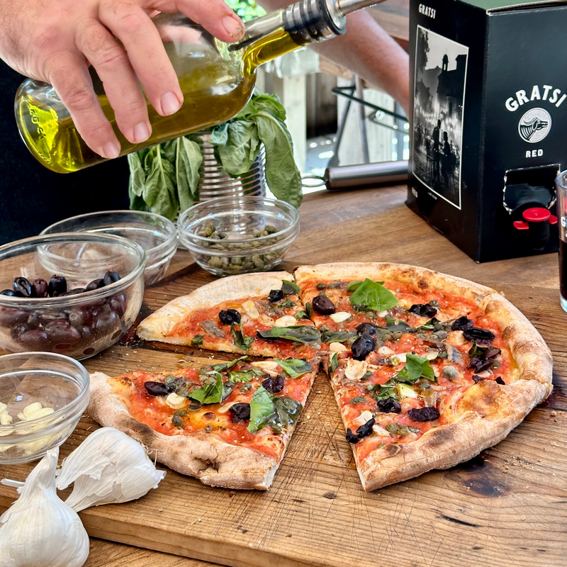 A hand pours olive oil over a sliced pizza on a wooden board, surrounded by ingredients including olives, garlic, capers, basil, and a box of red wine. Sunlight filters through the scene, creating a fresh outdoor ambiance.