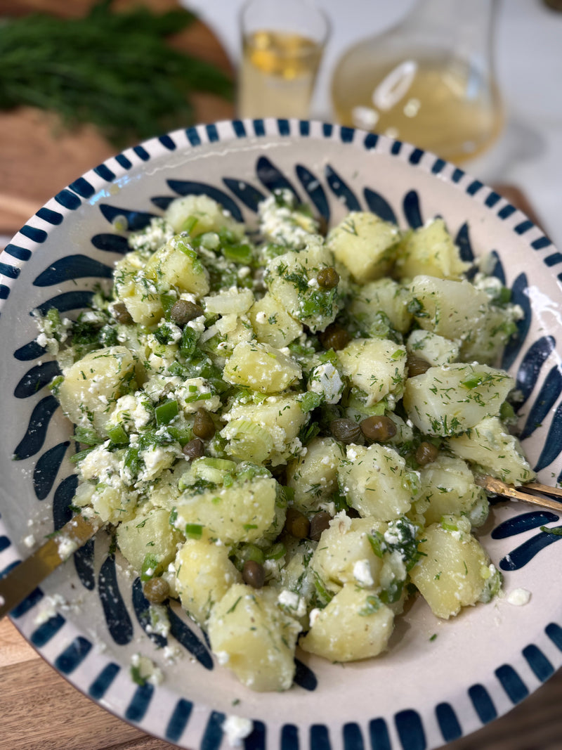 A bowl of potato salad with chopped herbs, capers, and crumbled feta cheese, served in a patterned dish. A glass and a carafe with a yellow beverage and fresh dill are in the blurred background.