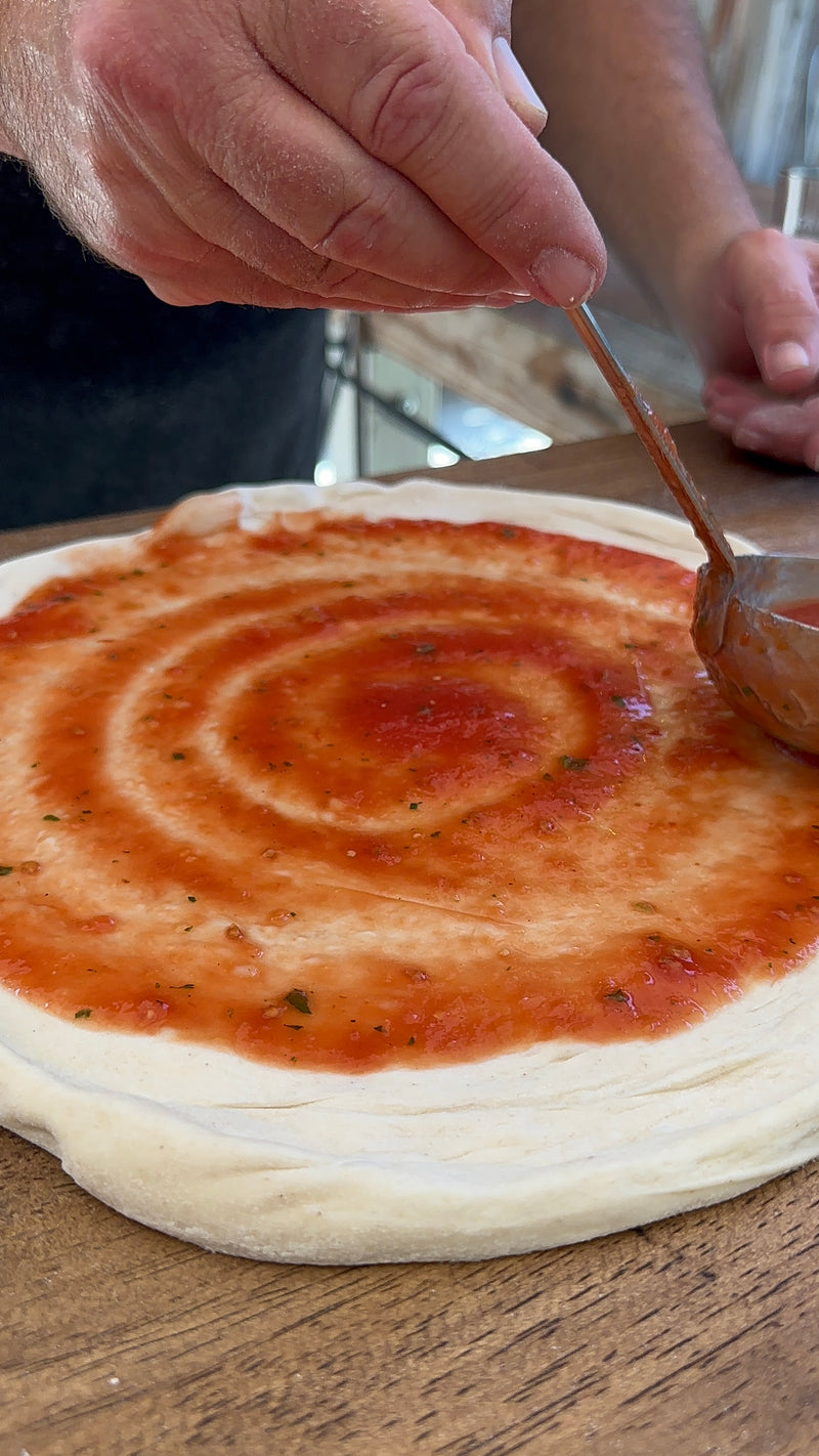 A close-up of a person spreading tomato sauce in a spiral pattern over a round pizza dough with a spoon on a wooden surface.