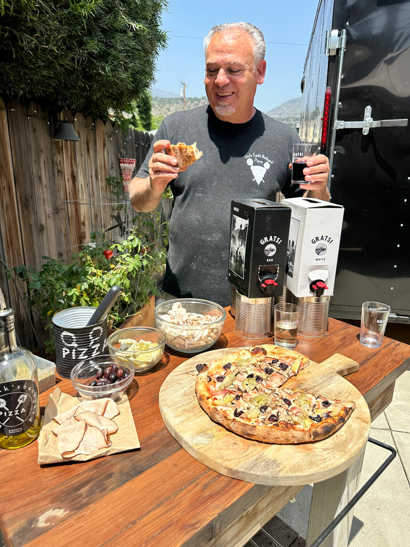 A man stands outdoors eating pizza and holding a glass of wine beside a table with a freshly baked pizza, deli meats, olives, mushrooms, olive oil, and boxed wine. A food truck and plants are visible in the background.