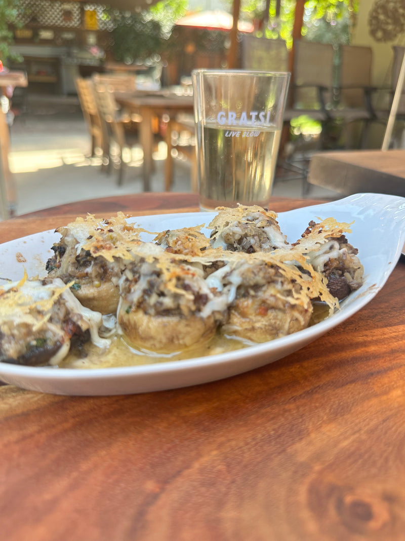 A white dish of stuffed mushrooms topped with melted cheese sits on a wooden table outdoors, with a glass of white wine and patio furniture visible in the background.