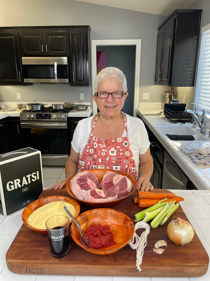 An older woman with short gray hair and glasses, wearing a red apron, stands in a kitchen behind a counter with pork, carrots, celery, onion, tomato paste, polenta, and a glass labeled GRATSI.
