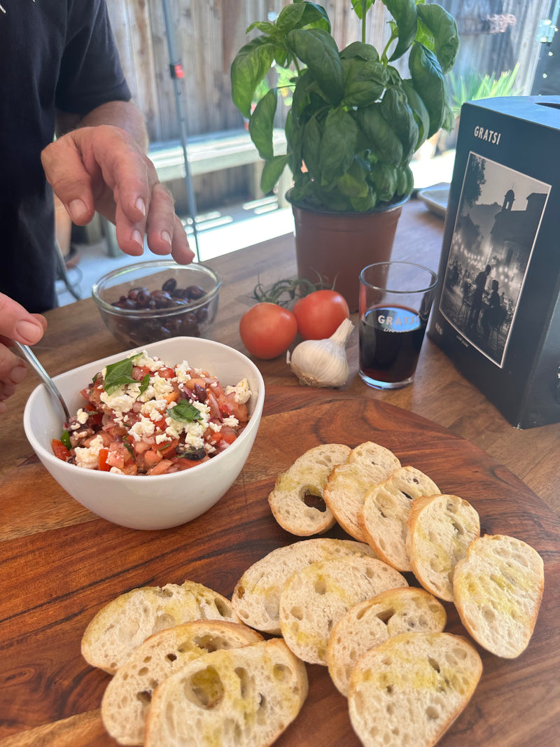 A bowl of salad with feta, tomatoes, and herbs sits on a wooden board with slices of toasted bread. Nearby are garlic, whole tomatoes, a basil plant, olives, a glass of red wine, and a Gratsi wine box.