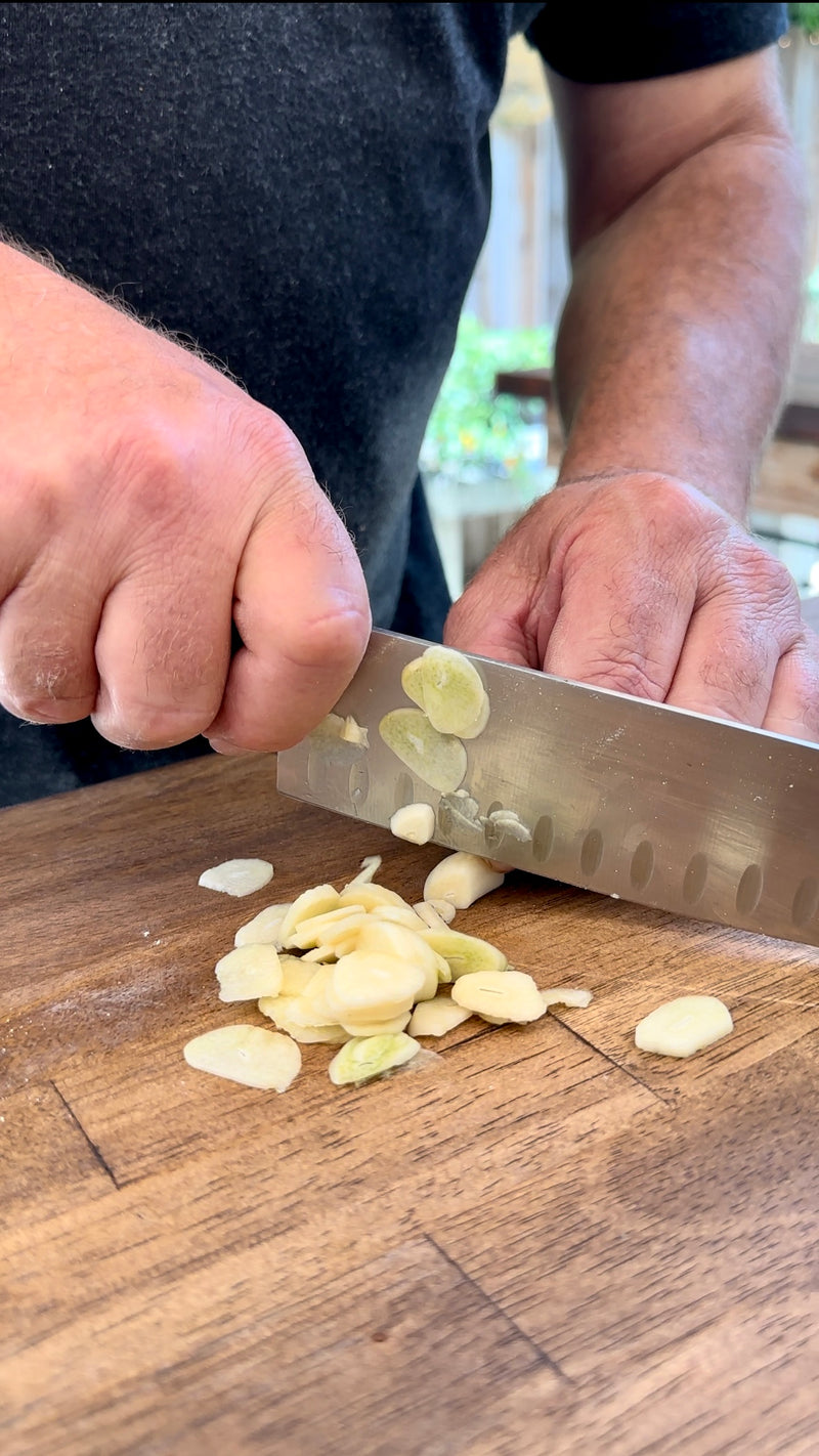 Close-up of a person slicing fresh garlic cloves with a large kitchen knife on a wooden cutting board. The thin garlic slices are gathered near the knife blade.