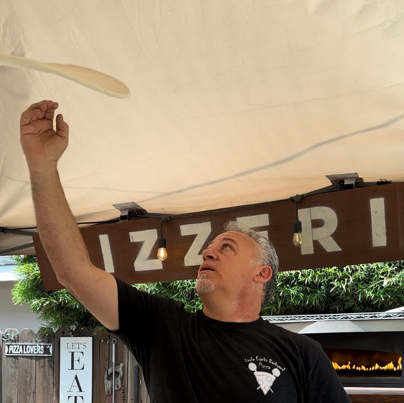 A man tossing pizza dough in the air under a tent, looking up at the dough. Behind him is a wooden sign with partially visible letters and string lights, with a pizza oven and greenery in the background.