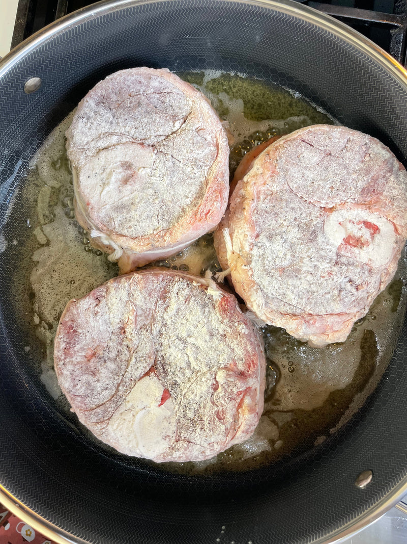Three floured beef shanks are sizzling in a skillet with oil, beginning to brown on the edges. The meat is arranged in a single layer in a nonstick pan on a stovetop.
