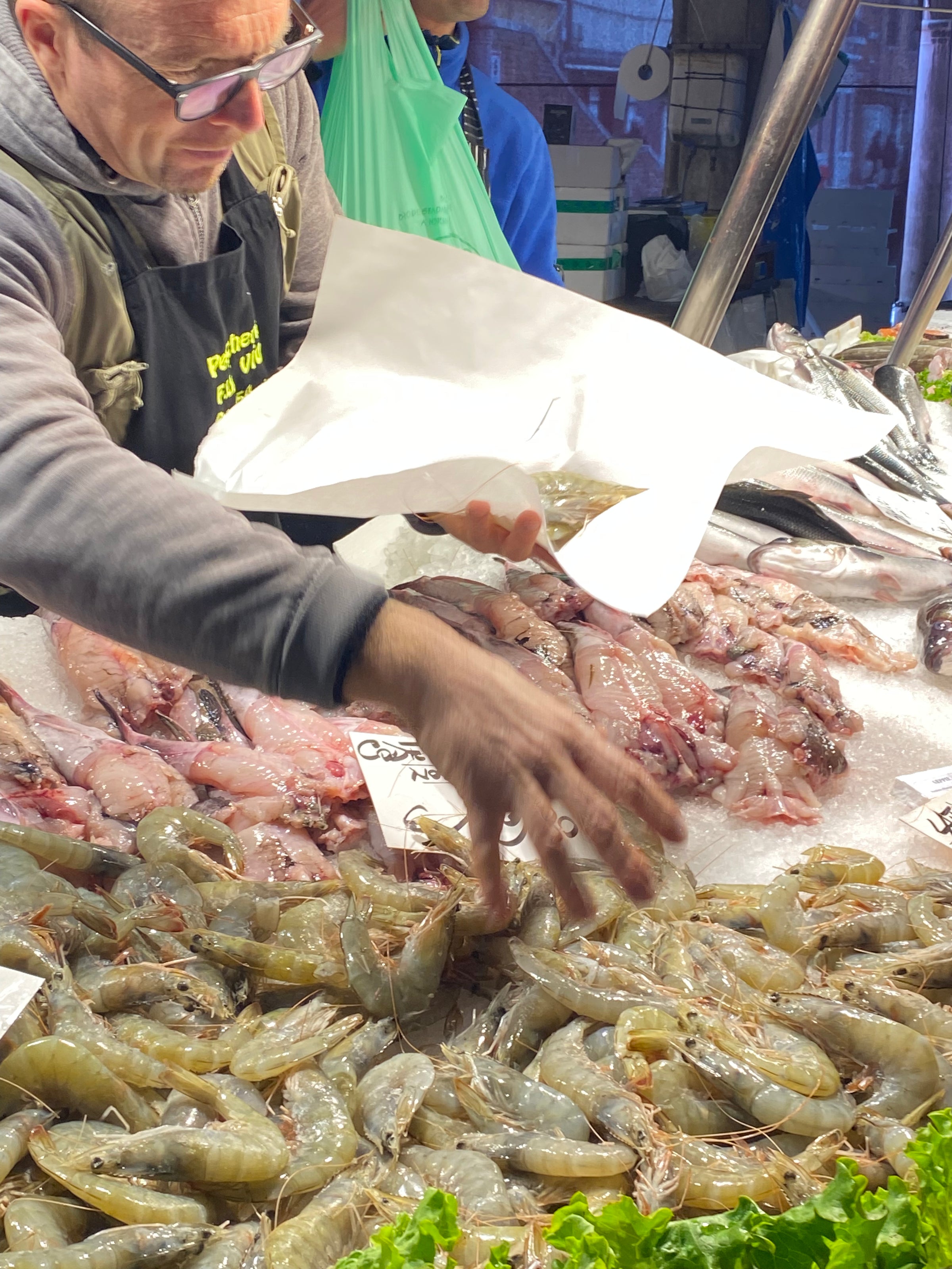 A seafood vendor wearing glasses selects prawns from a display of fresh seafood on ice, including fish and shrimp, at an outdoor market stall.