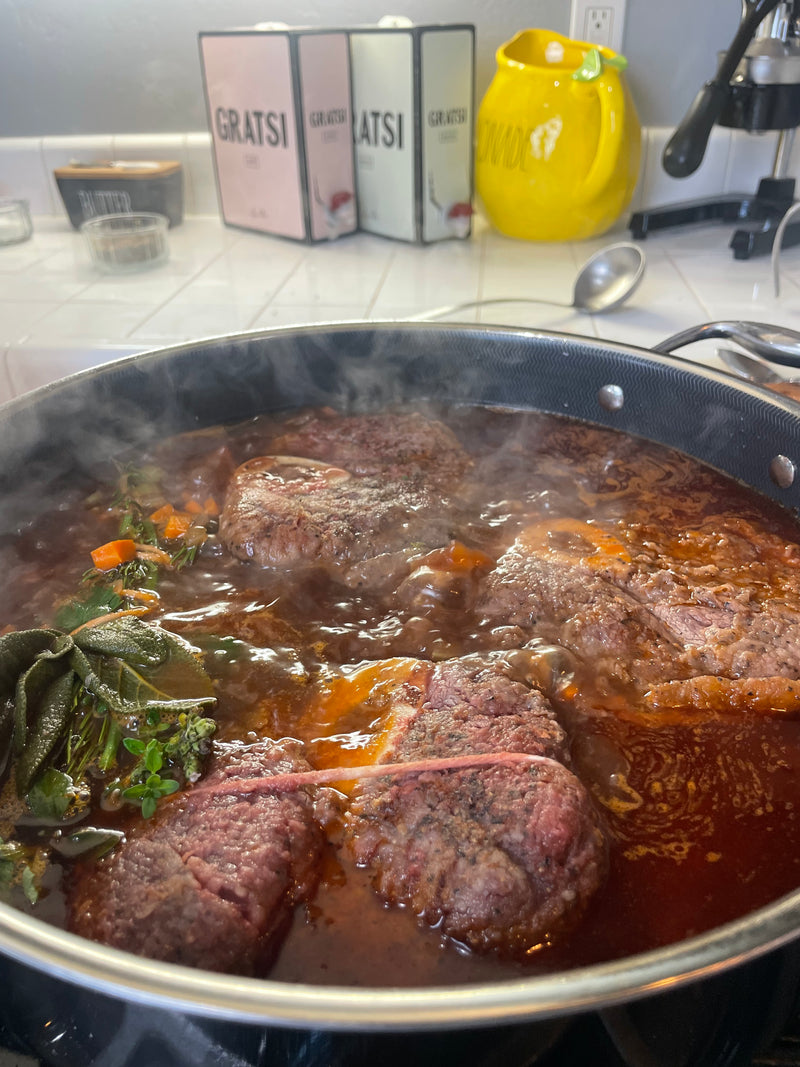 Close-up of beef simmering in a rich, red sauce with herbs and vegetables in a black pot on a stovetop. Steam rises from the pot, and kitchen items are visible in the background.