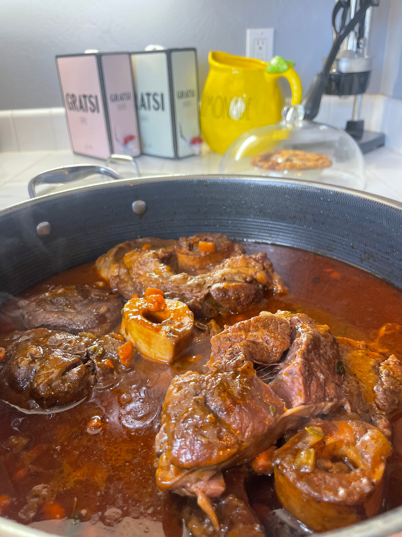 A pot of braised beef shanks in rich red sauce simmers on a stove. In the background, yellow and white kitchen containers labeled “GRATSI” and “HONEY” sit on a white countertop.