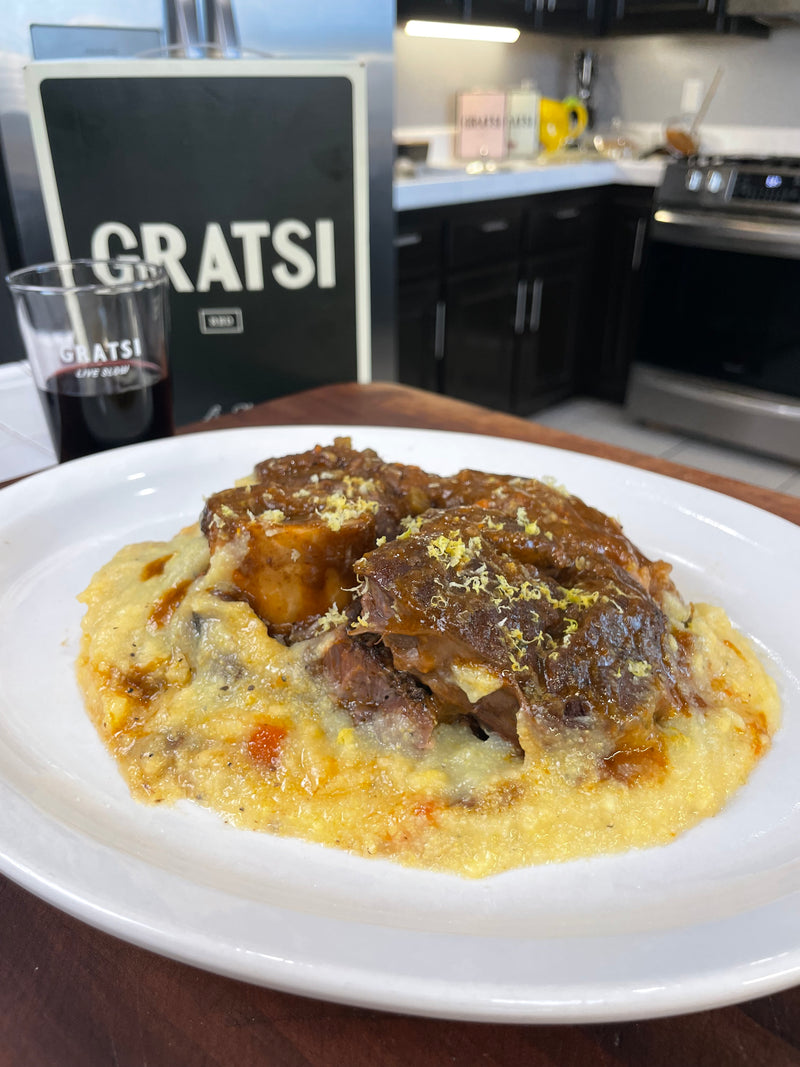 A plate of braised meat served over creamy polenta is on a wooden counter. In the background, there’s a glass of red wine and a menu or sign that reads “GRATSI,” with a kitchen visible behind.
