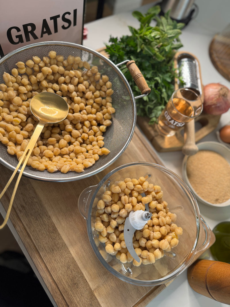 Chickpeas are draining in a colander with a gold spoon, while more chickpeas are in a food processor. Fresh parsley, onion, olive oil, a jar labeled GRATSI, and other ingredients are arranged on a wooden surface.