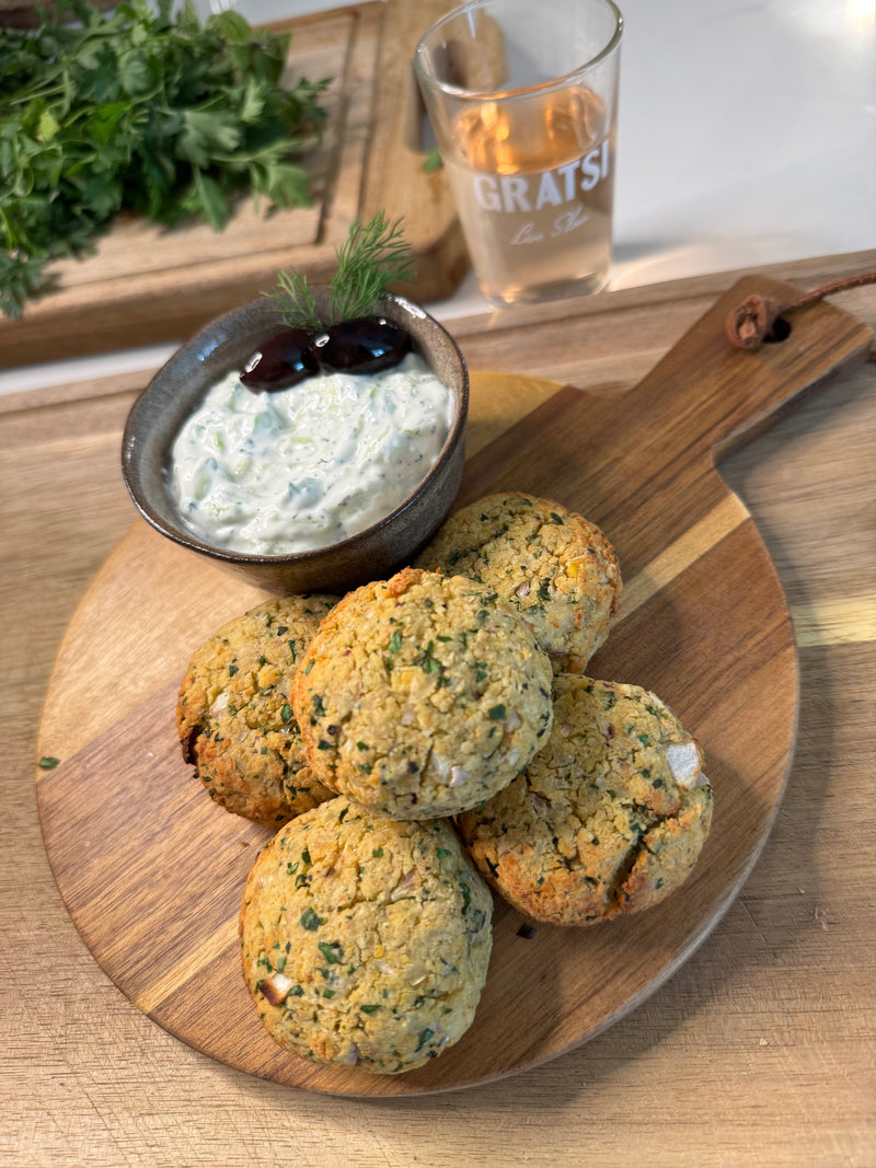Five baked falafel patties on a wooden board, served with a bowl of creamy tzatziki sauce garnished with olives and dill. A glass of rosé and fresh herbs are in the background.
