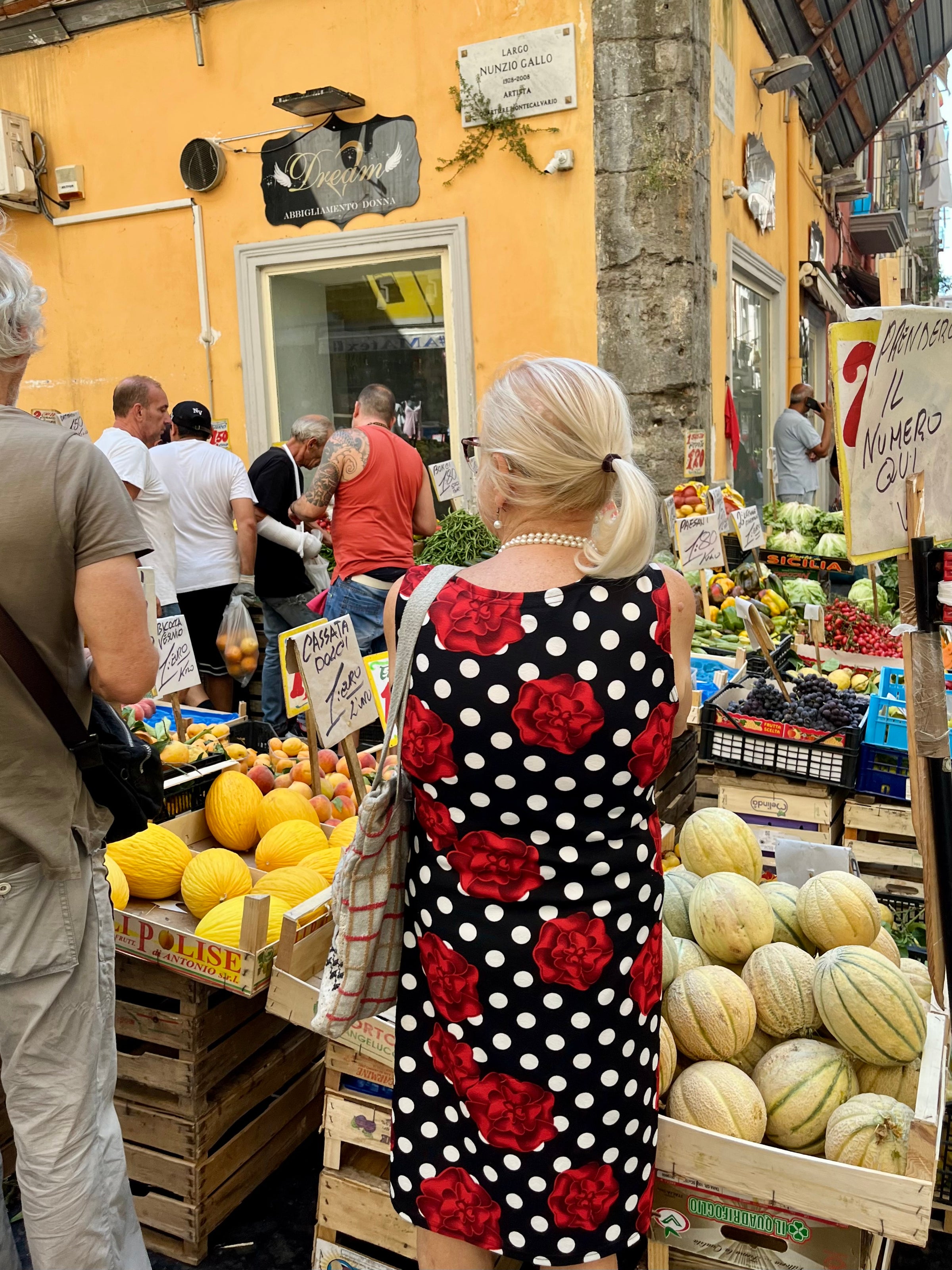 A woman in a black dress with red roses and white polka dots stands at an outdoor market stall filled with melons and fresh produce. Other shoppers and vendors are visible in the busy scene.