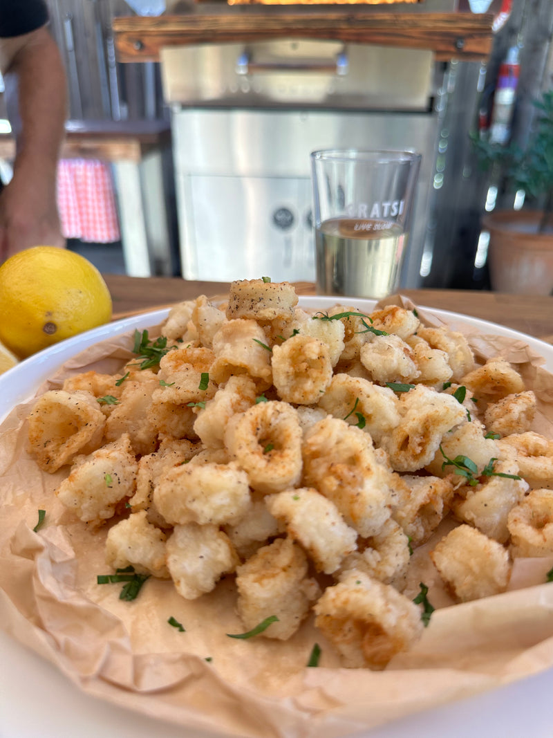 A plate of golden, crispy fried calamari garnished with chopped herbs sits on parchment paper. A sliced lemon and a glass of white wine are in the background, with a blurred kitchen scene behind.