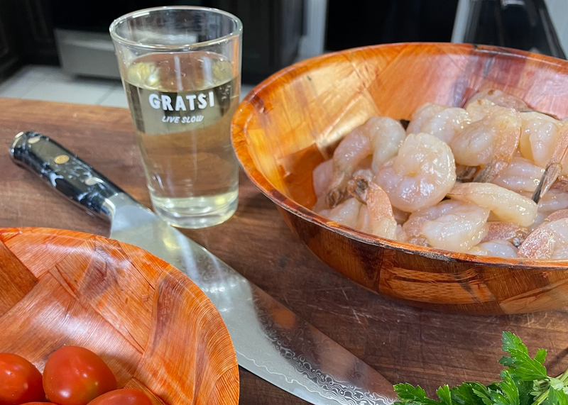 A wooden bowl filled with raw shrimp sits next to a chef’s knife, a glass with white wine labeled “GRATSI LIVE SLOW,” a bowl of cherry tomatoes, and some fresh parsley on a kitchen counter.