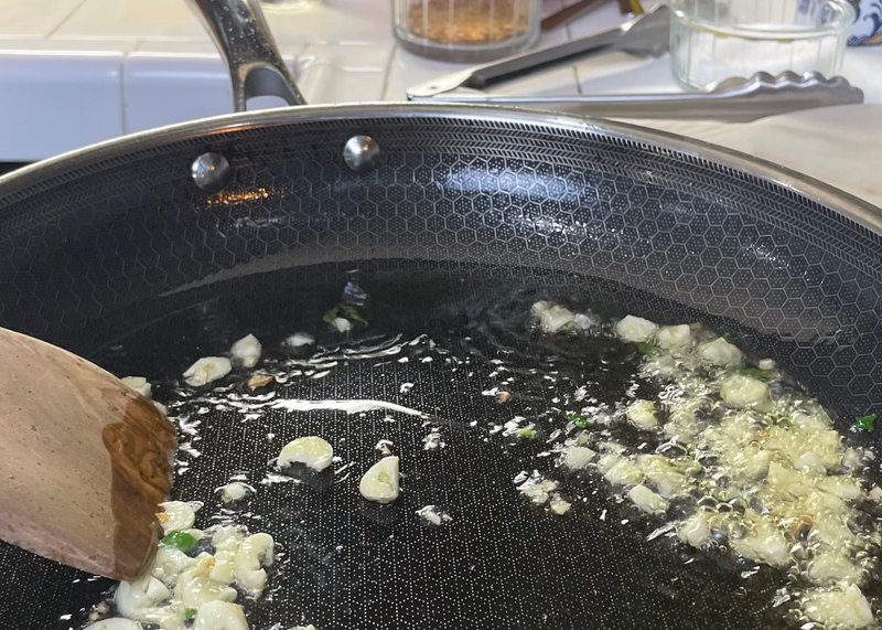 A close-up of a frying pan with chopped garlic and green herbs sizzling in oil. A wooden spatula is stirring the mixture. Kitchen utensils and spice containers are visible in the background.
