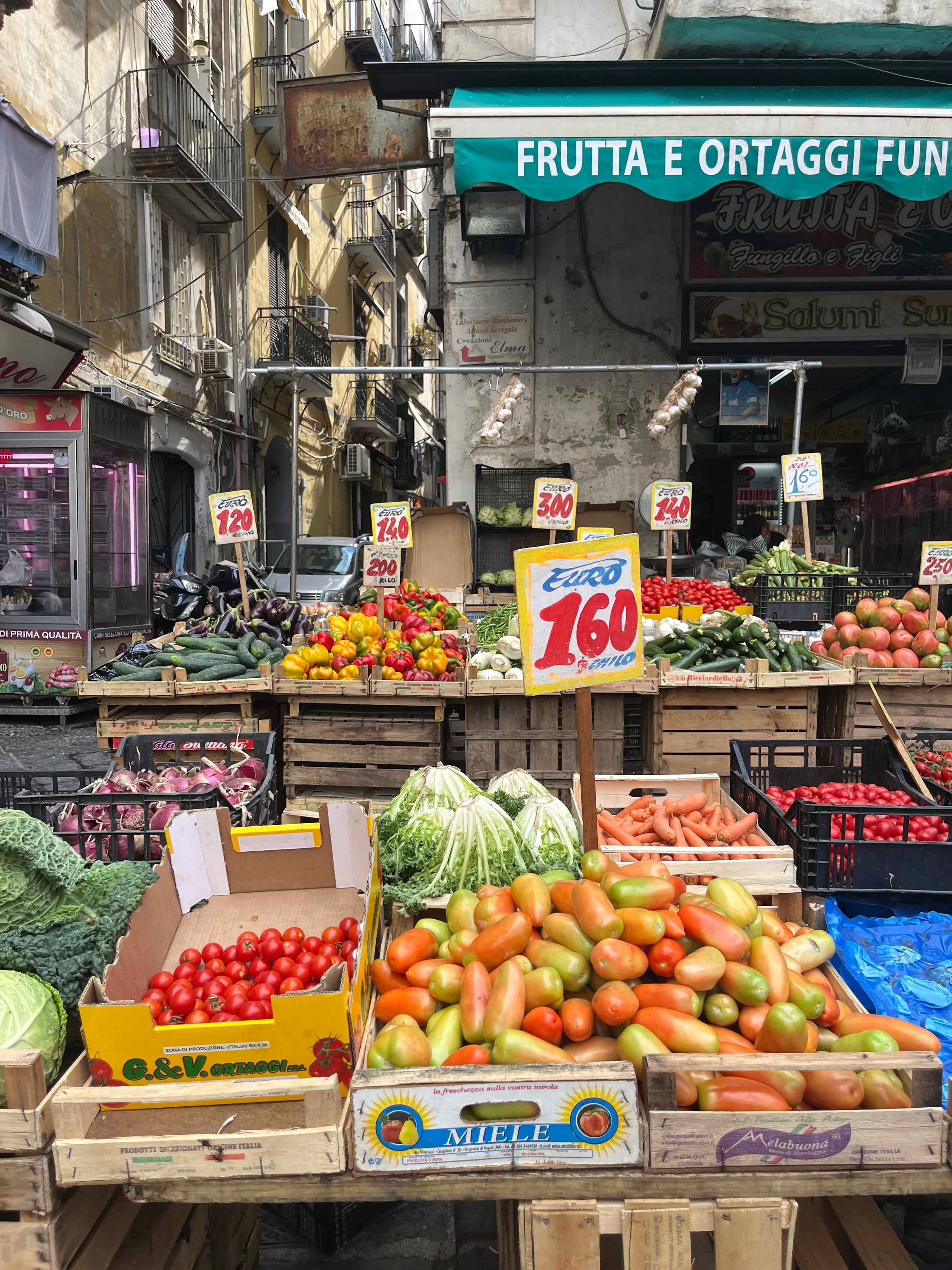 Outdoor market stand with crates of fresh vegetables including tomatoes, carrots, cabbage, and green beans, under a green awning reading FRUTTA E ORTAGGI. Price signs are displayed on the produce.