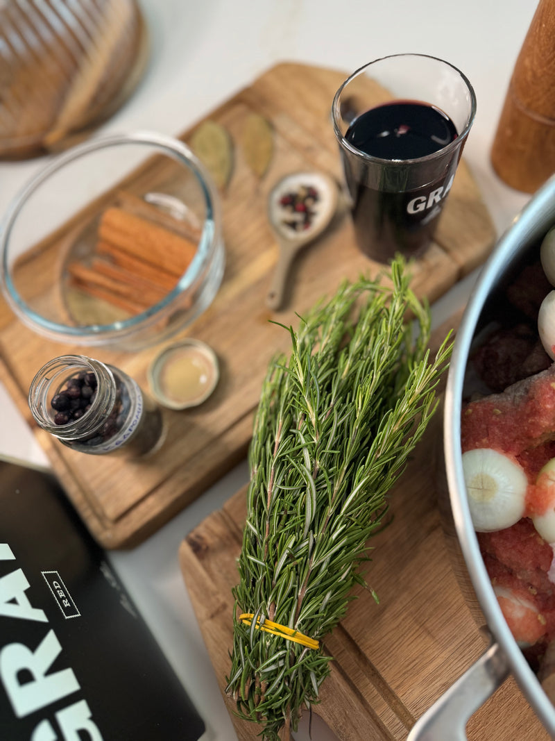 A bundle of fresh rosemary on a wooden cutting board beside a pot with onions and meat, a glass of red wine, spices in jars, bay leaves, cinnamon sticks, and peppercorns.