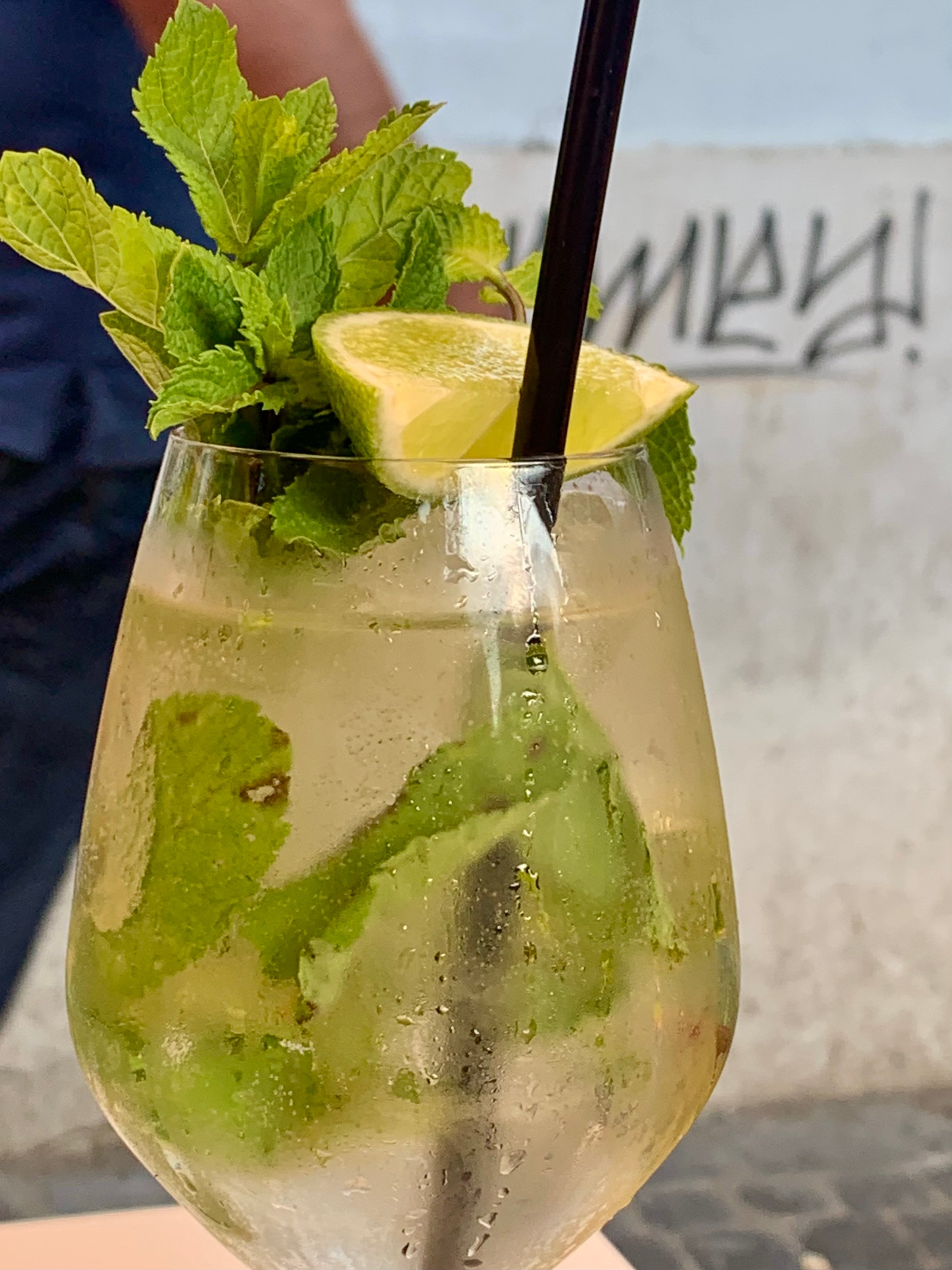 A close-up of a glass filled with a light-colored cocktail, garnished with fresh mint leaves, a lime wedge, and a black straw. The background shows a blurred wall with graffiti.