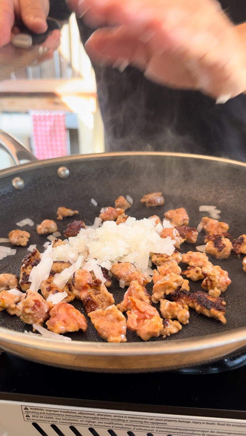 Chopped onions and browned sausage pieces cooking in a frying pan, with a persons hand visible above the pan. Steam rises from the sizzling ingredients.