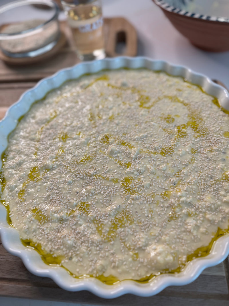 A round baking dish filled with an unbaked dough topped with sesame seeds and drizzled with olive oil sits on a wooden surface, with bowls and ingredients visible in the background.