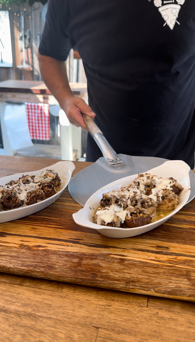 A person serves a baked dish topped with white sauce from a white oval dish onto a plate, with another identical dish beside it on a wooden table.