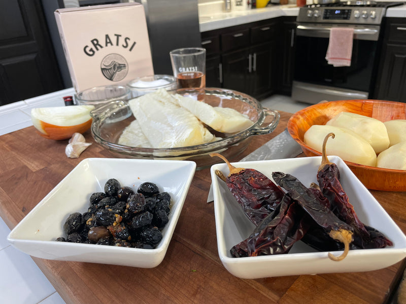 A kitchen counter with white bowls containing black olives and dried red chilies, a glass dish with white fish fillets, peeled potatoes, onion, and a Gratsi wine box and glass in the background.