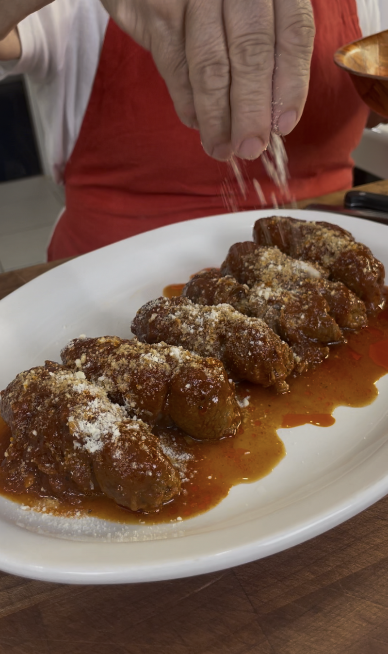 A person sprinkles grated cheese over a row of meatballs covered in tomato sauce on a white plate. The person is wearing a red apron.