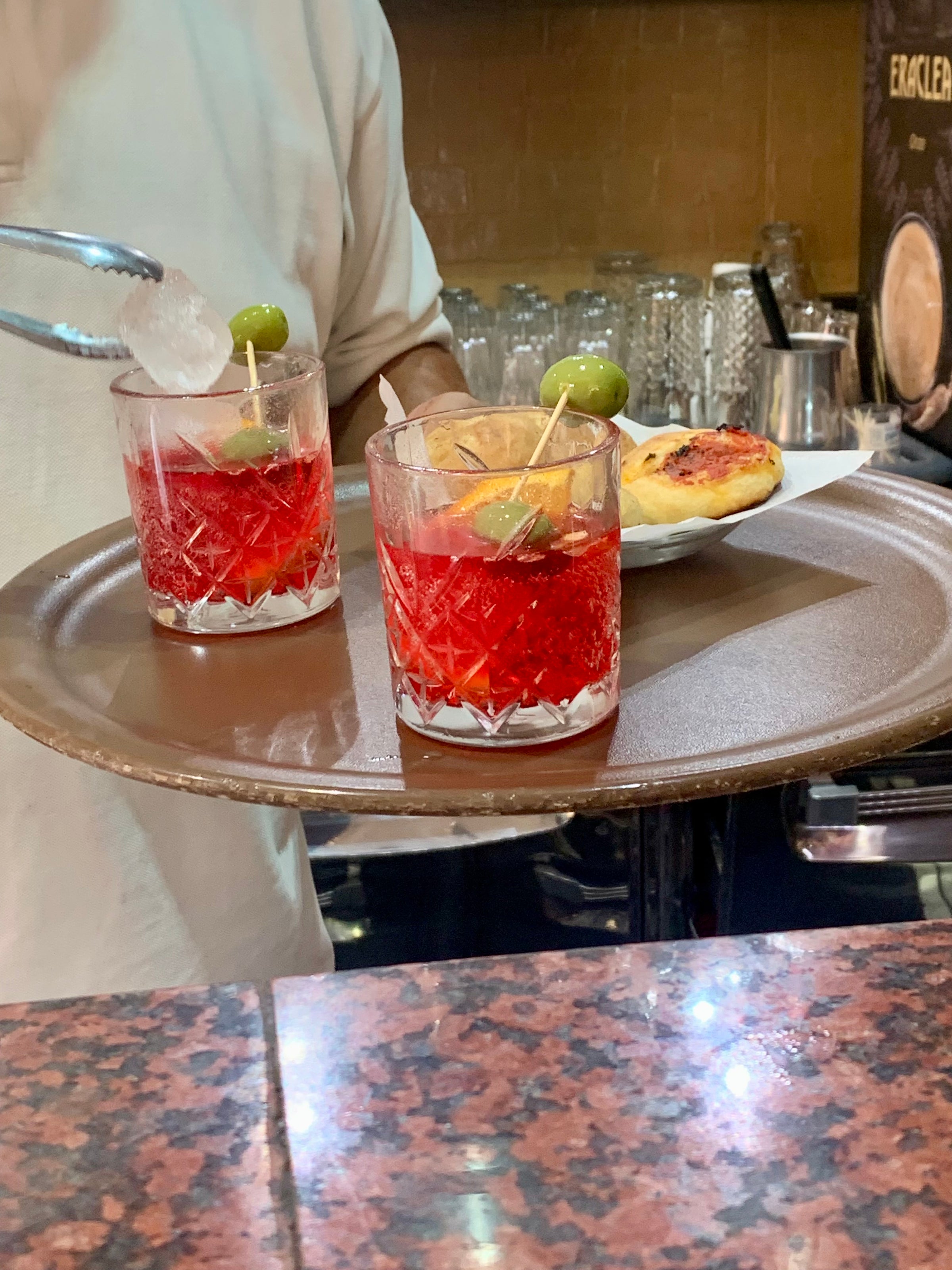 A server holds a tray with two red cocktails garnished with green olives and ice, and a small savory pastry beside them, on a marble countertop in a bar or café setting.