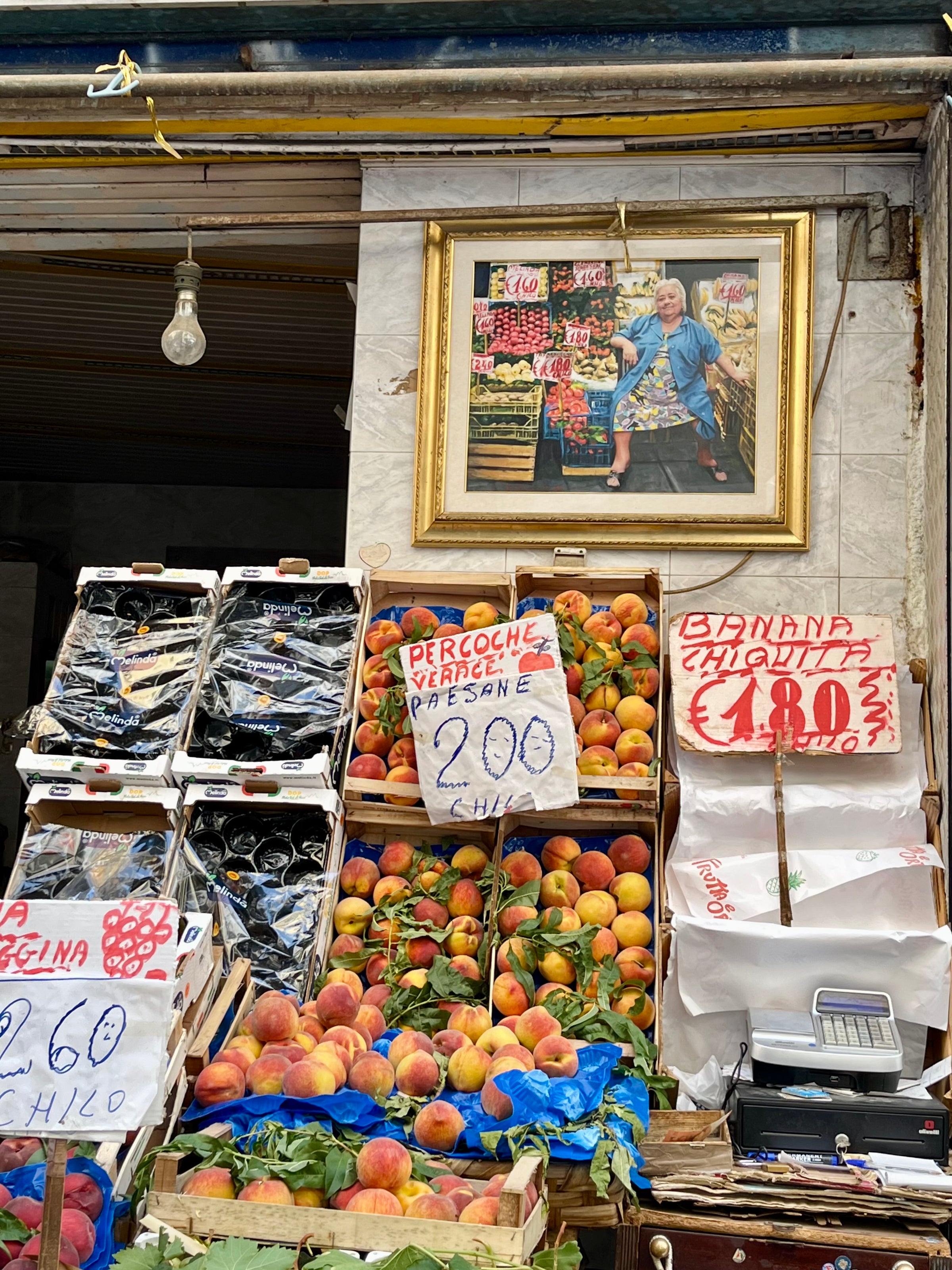 An outdoor fruit stand displays crates of peaches and cherries with handwritten price signs. Above the fruit, a framed photo of an elderly woman sitting and smiling hangs on the wall. A hanging lightbulb is visible.